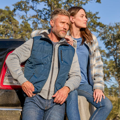 Man wearing the Free Country Men's FreeCycle® Stimson Puffer Vest in navy, featuring quilted design, REPREVE® poly fill, and a vertical zip, layered over a burgundy long-sleeve shirt with dark jeans.