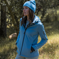 Woman wearing Free Country Women's StormTech Super Softshell® Jacket in blue with a full front zipper opening, attached hood with adjustable cord, lower pockets, soft butter pile fabric lining, and water resistant and wind resistant stretch performance fabric. light grey background. Best women’s jacket for fall weather.