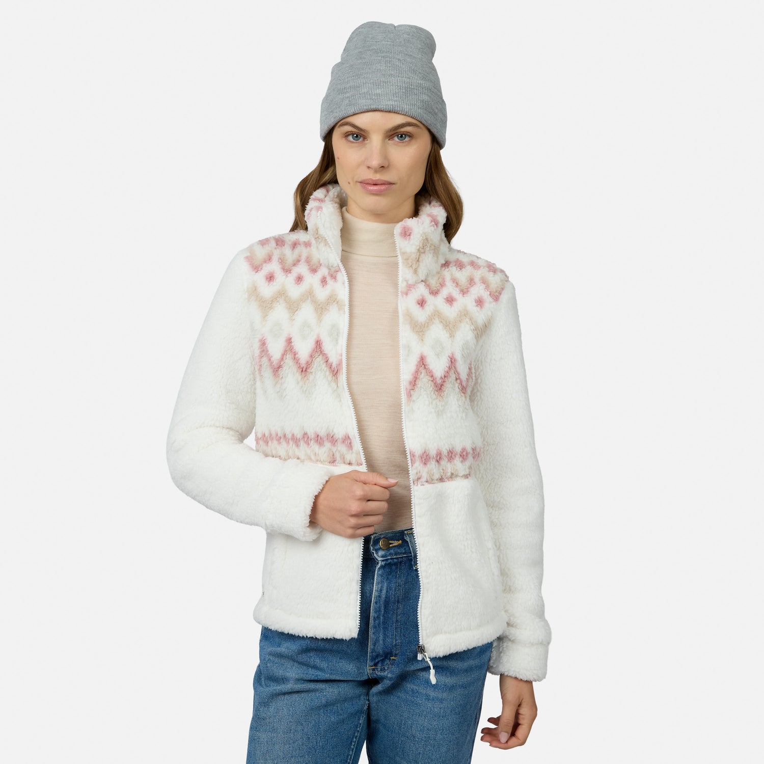 Woman in a gray beanie, light pink turtleneck, blue jeans, and the Free Country Women’s Sierra Butter Pile® Zip Up Jacket with pink and beige patterns stands against a white background. This stylish jacket is lightweight and cozy.