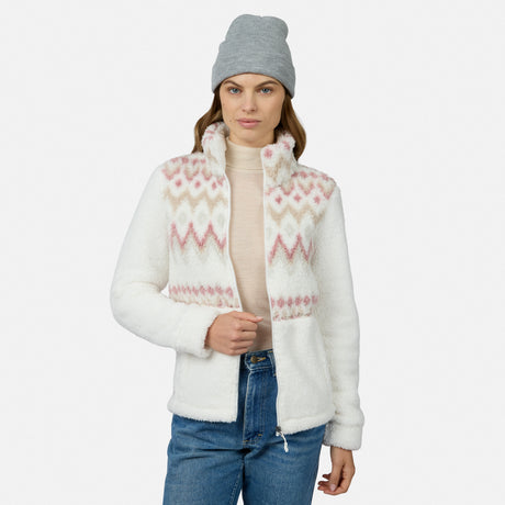 Woman in a gray beanie, light pink turtleneck, blue jeans, and the Free Country Women’s Sierra Butter Pile® Zip Up Jacket with pink and beige patterns stands against a white background. This stylish jacket is lightweight and cozy.