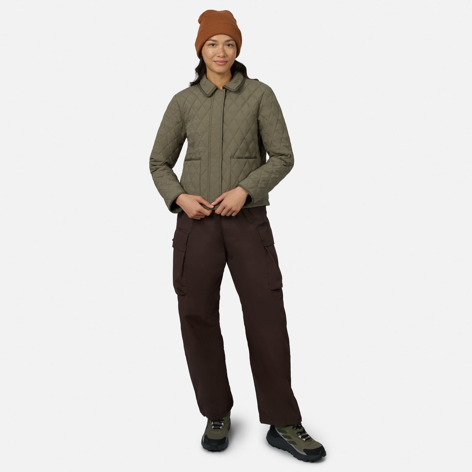 [Model is wearing size S.] A model wearing a quilted olive green jacket, dark brown cargo pants, a rust-colored beanie, and hiking boots, standing against a light gray studio background.