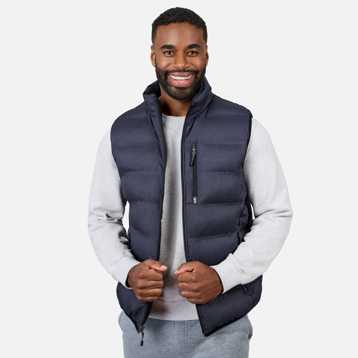 A man smiles while wearing the Free Country Men’s Haven Stitchless Reversible Vest in navy blue, which provides lightweight warmth, layered over a light gray sweatshirt and matching gray pants, standing against a plain white background.
