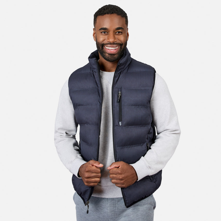 A man smiles while wearing the Free Country Men’s Haven Stitchless Reversible Vest in navy blue, which provides lightweight warmth, layered over a light gray sweatshirt and matching gray pants, standing against a plain white background.