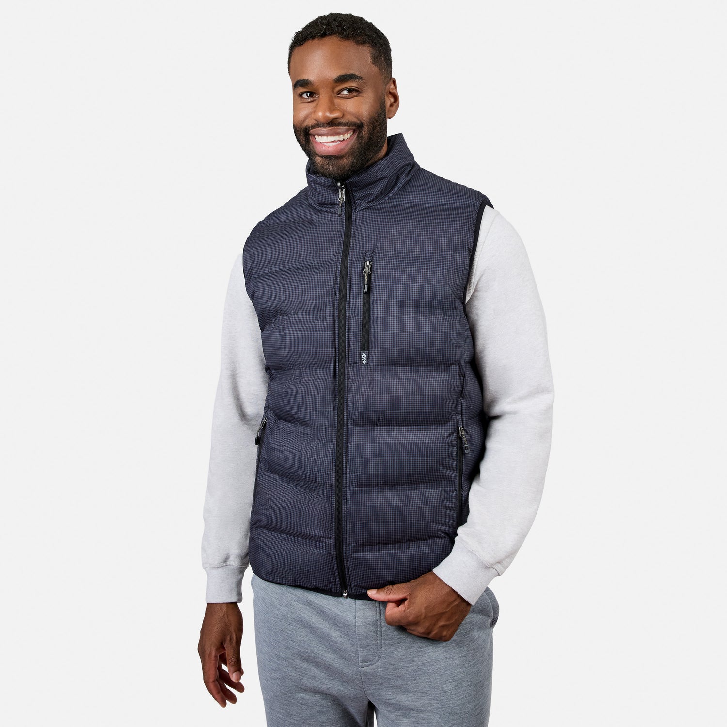 A man smiles while wearing the Free Country Men’s Haven Stitchless Reversible Vest in navy, featuring a lightweight puffer exterior and cozy sherpa reverse, layered over a light gray sweatshirt and gray sweatpants as he stands against a plain white background.