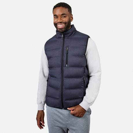 A man smiles while wearing the Free Country Men’s Haven Stitchless Reversible Vest in navy, featuring a lightweight puffer exterior and cozy sherpa reverse, layered over a light gray sweatshirt and gray sweatpants as he stands against a plain white background.