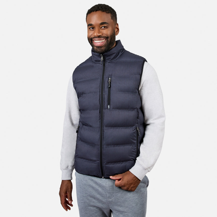 A man smiles while wearing the Free Country Men’s Haven Stitchless Reversible Vest in navy, featuring a lightweight puffer exterior and cozy sherpa reverse, layered over a light gray sweatshirt and gray sweatpants as he stands against a plain white background.