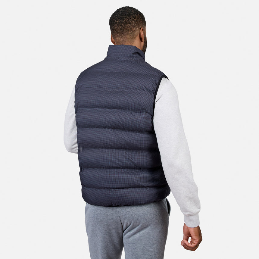 A man stands facing away against a plain white background, wearing the Free Country Men’s Haven Stitchless Reversible Vest in navy blue with sherpa reverse over a light gray sweatshirt and gray sweatpants, offering lightweight warmth and versatile style.