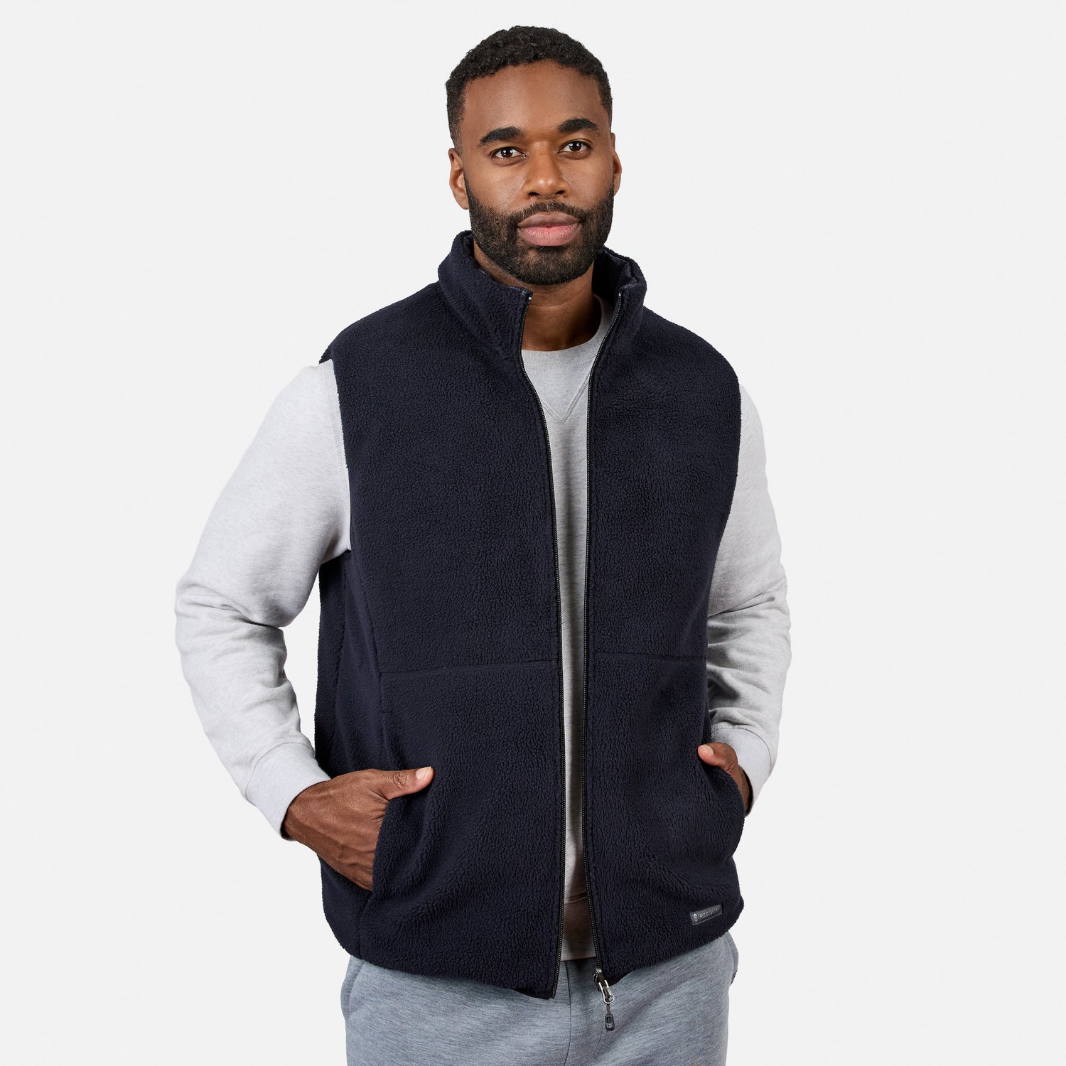 A man wearing the Free Country Men’s Haven Stitchless Reversible Vest in dark navy with its sherpa side reversed, layered over a light gray sweatshirt and gray pants, stands with his hands in his vest pockets against a plain white background, highlighting the vest’s lightweight warmth and versatile style.