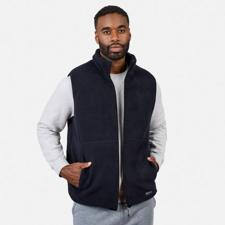 A man wearing the Free Country Men’s Haven Stitchless Reversible Vest in dark navy with its sherpa side reversed, layered over a light gray sweatshirt and gray pants, stands with his hands in his vest pockets against a plain white background, highlighting the vest’s lightweight warmth and versatile style.