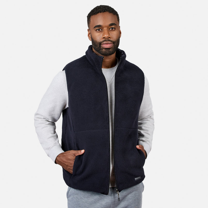 A man wearing the Free Country Men’s Haven Stitchless Reversible Vest in dark navy with its sherpa side reversed, layered over a light gray sweatshirt and gray pants, stands with his hands in his vest pockets against a plain white background, highlighting the vest’s lightweight warmth and versatile style.