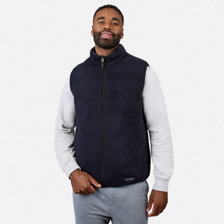 A man stands against a light gray background, wearing the Free Country Men’s Haven Stitchless Reversible Vest in black with sherpa reverse over a light gray sweatshirt and gray pants. He is looking slightly to the side and smiling with one hand in his pocket, enjoying the lightweight warmth and casual comfort of this versatile vest.