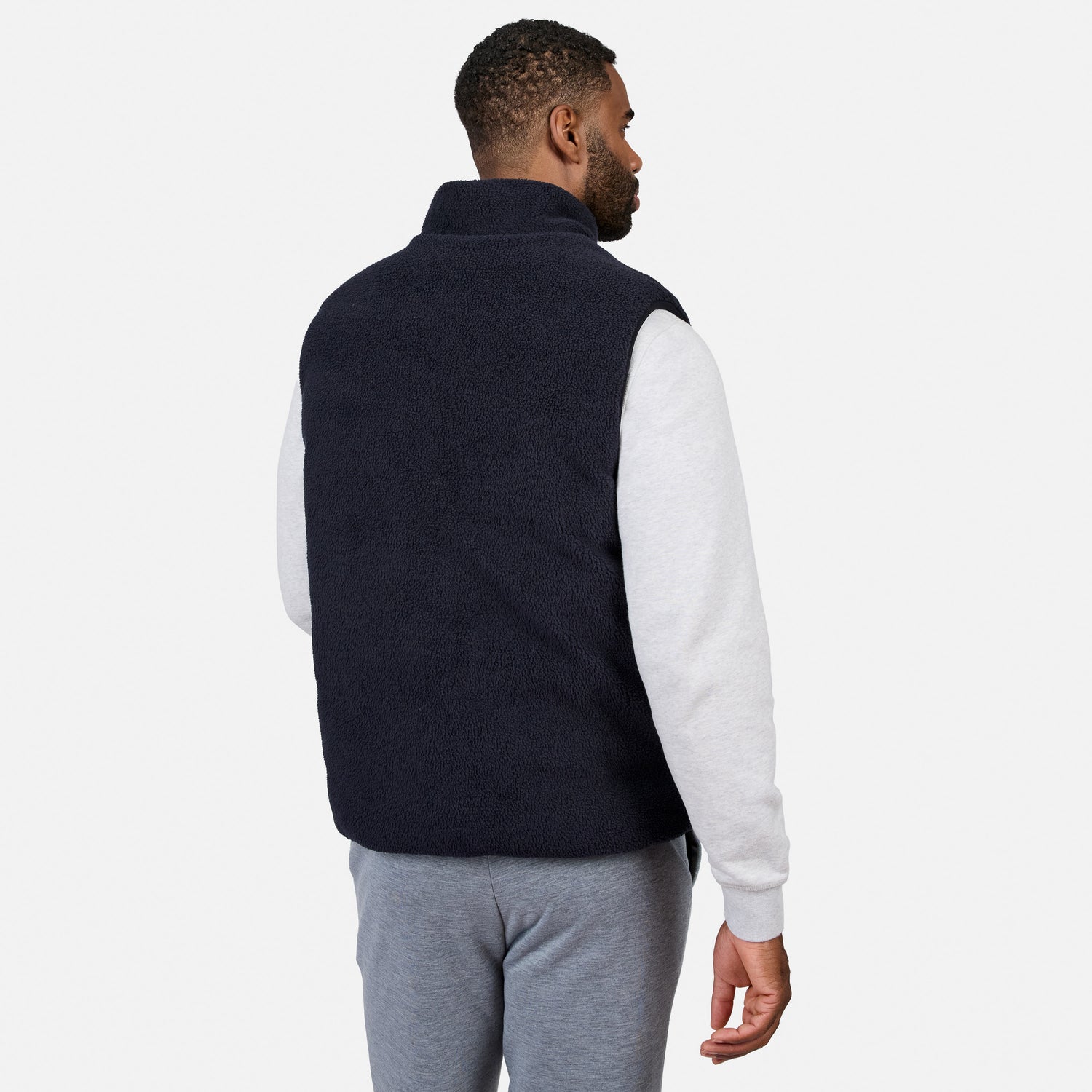 A man stands facing away, wearing the Free Country Men’s Haven Stitchless Reversible Vest in navy over a light gray sweatshirt and gray sweatpants, set against a plain white background.