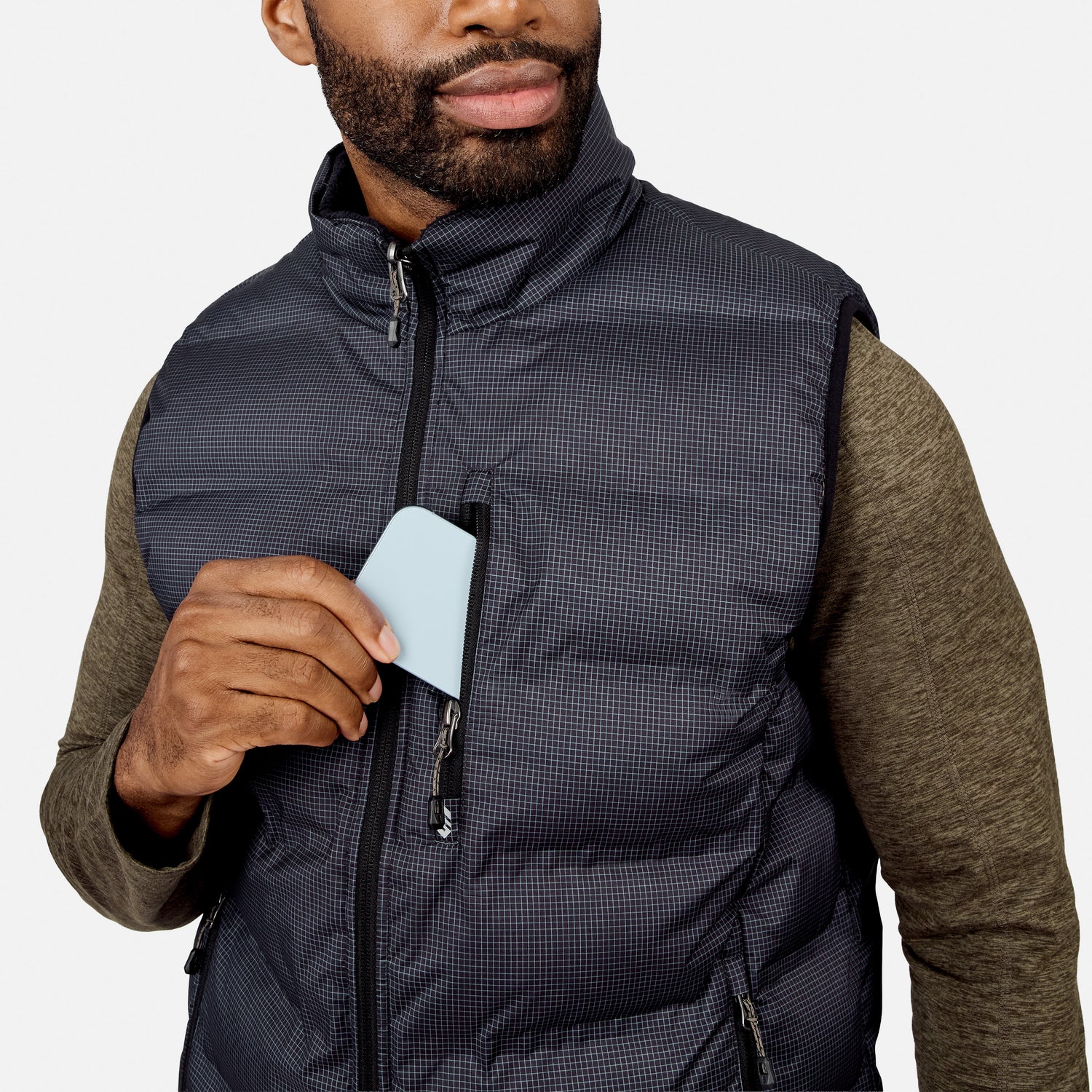 Wearing the Free Country Men’s Haven Stitchless Reversible Vest over a green shirt, a man slips a light blue card into the vest’s chest pocket, appreciating its lightweight warmth.