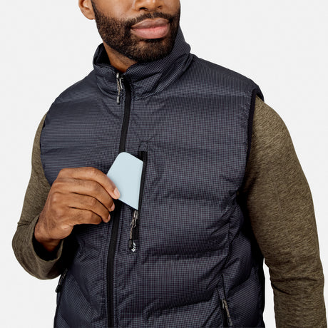 Wearing the Free Country Men’s Haven Stitchless Reversible Vest over a green shirt, a man slips a light blue card into the vest’s chest pocket, appreciating its lightweight warmth.