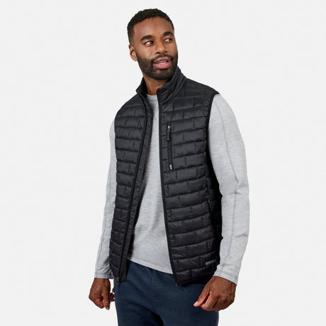 [Model is wearing size M.] A man wearing a black quilted zip-front vest over a light gray long-sleeve shirt and dark blue jeans, photographed against a clean light gray studio background.