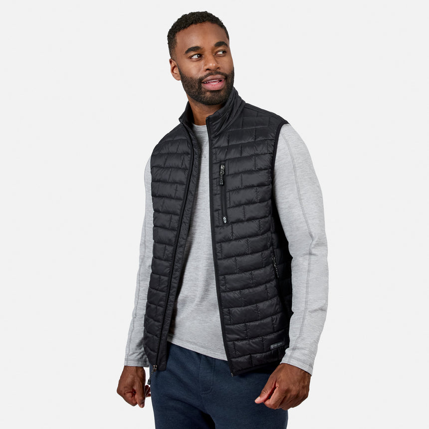 [Model is wearing size M.] A man wearing a black quilted zip-front vest over a light gray long-sleeve shirt and dark blue jeans, photographed against a clean light gray studio background.