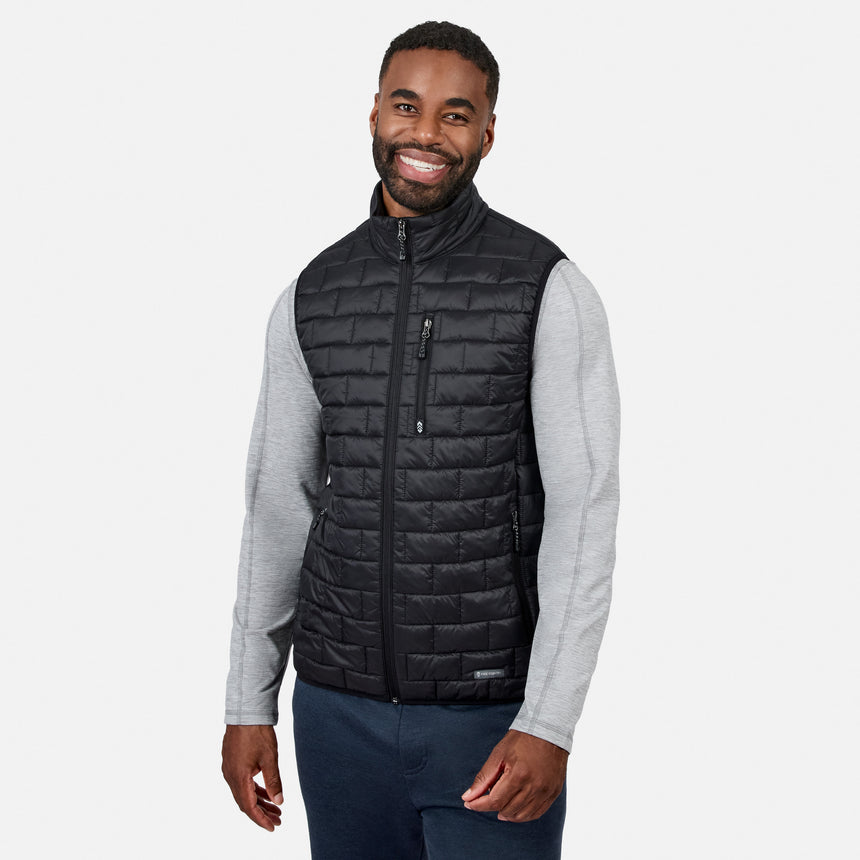 [Model is wearing size M.] A smiling man wearing a black quilted zip-front puffer vest over a light gray long-sleeve shirt and navy pants, photographed against a clean light gray studio background.
