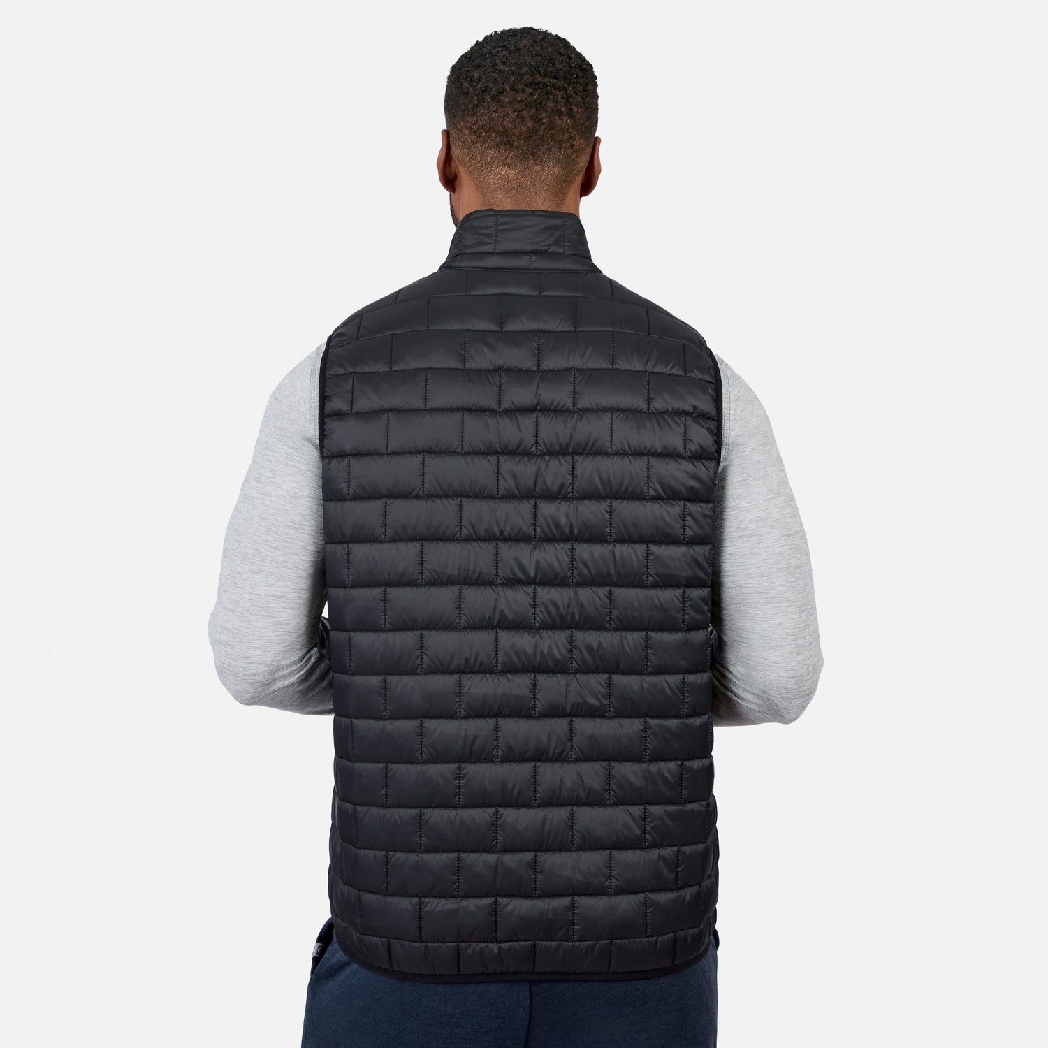 [Model is wearing size M.] A man viewed from behind wearing a black quilted puffer vest over a light gray long-sleeve shirt and dark blue pants, against a plain light gray studio background.