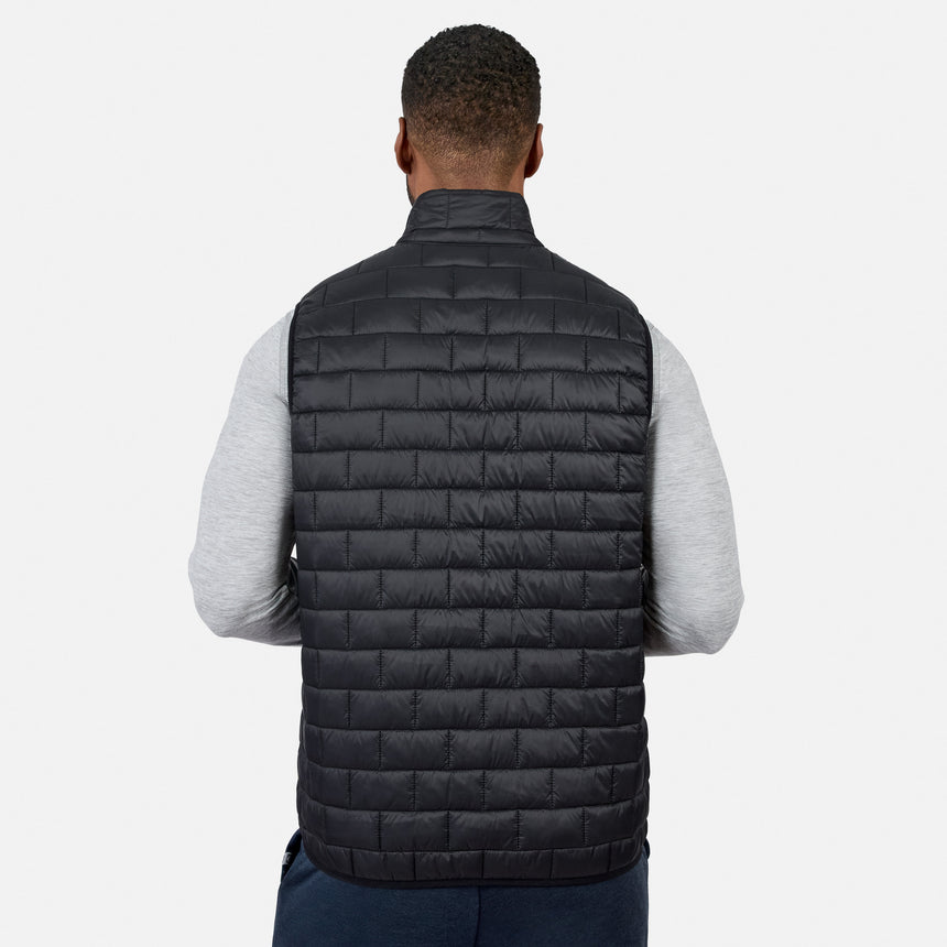 [Model is wearing size M.] A man viewed from behind wearing a black quilted puffer vest over a light gray long-sleeve shirt and dark blue pants, against a plain light gray studio background.