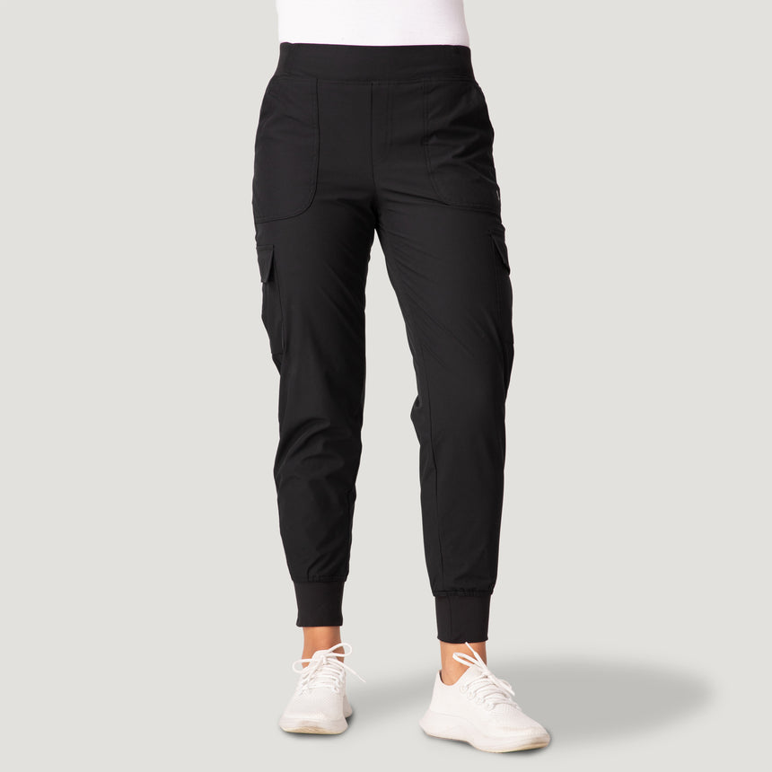 A woman wears Free Country’s Women’s Get Out There Fleece Lined Jogger in black, paired with a white top and white sneakers, standing facing forward against a light gray background.
