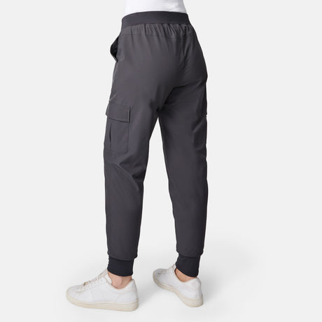 Woman models Free Country Women’s Get Out There Fleece Lined Jogger in dark gray with large side pocket, ribbed cuffs, and relaxed fit, paired with white sneakers. Shown from the back on a white background; pants are water-resistant cargo joggers.