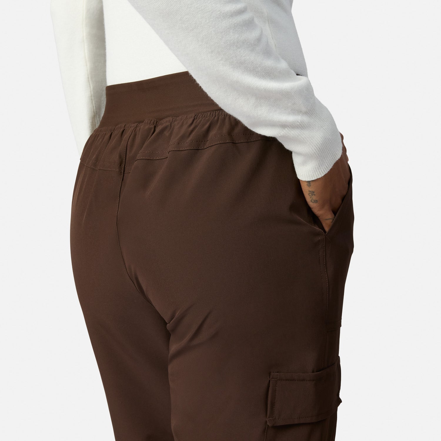 A person shown from the back with one hand in their pocket, wearing brown Free Country Women’s Get Out There Fleece Lined Jogger pants and a white long-sleeve top.