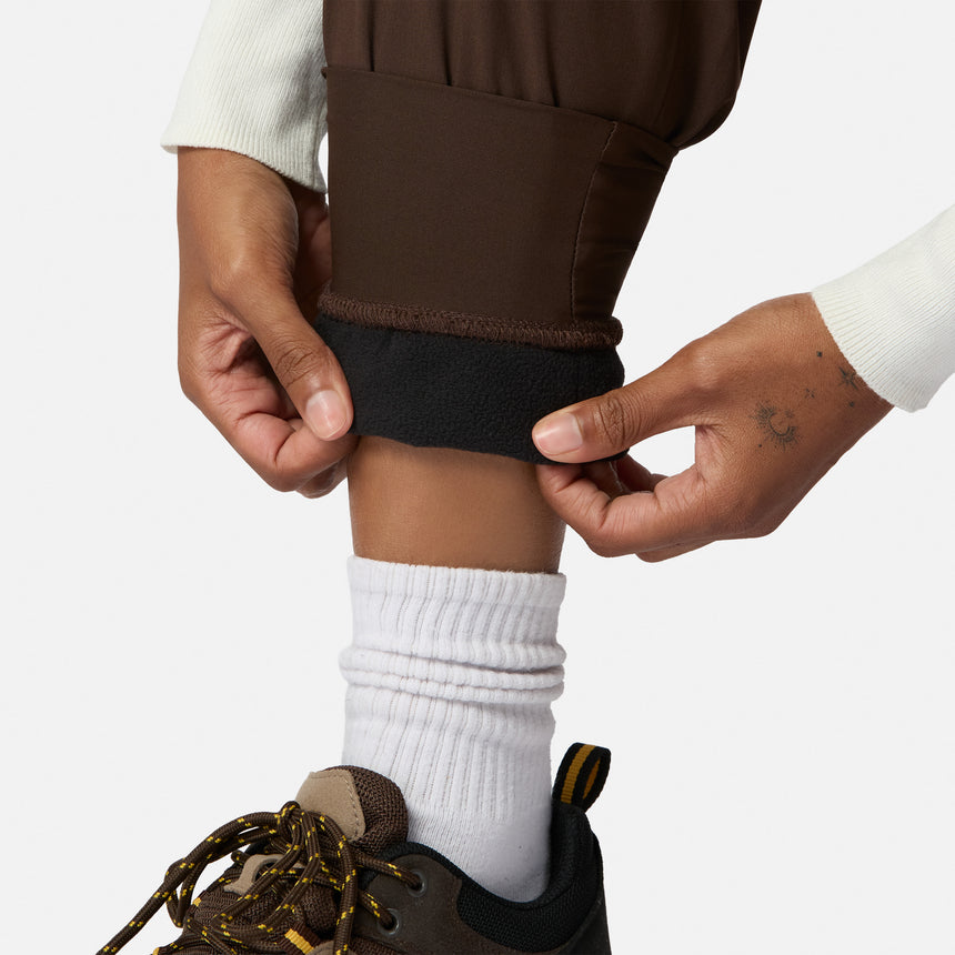 A person rolls up the hem of Free Country Women’s Get Out There Fleece Lined Jogger, exposing black fleece. They wear white socks, brown hiking shoes with yellow laces, a small hand tattoo is visible, all set against a plain light background.