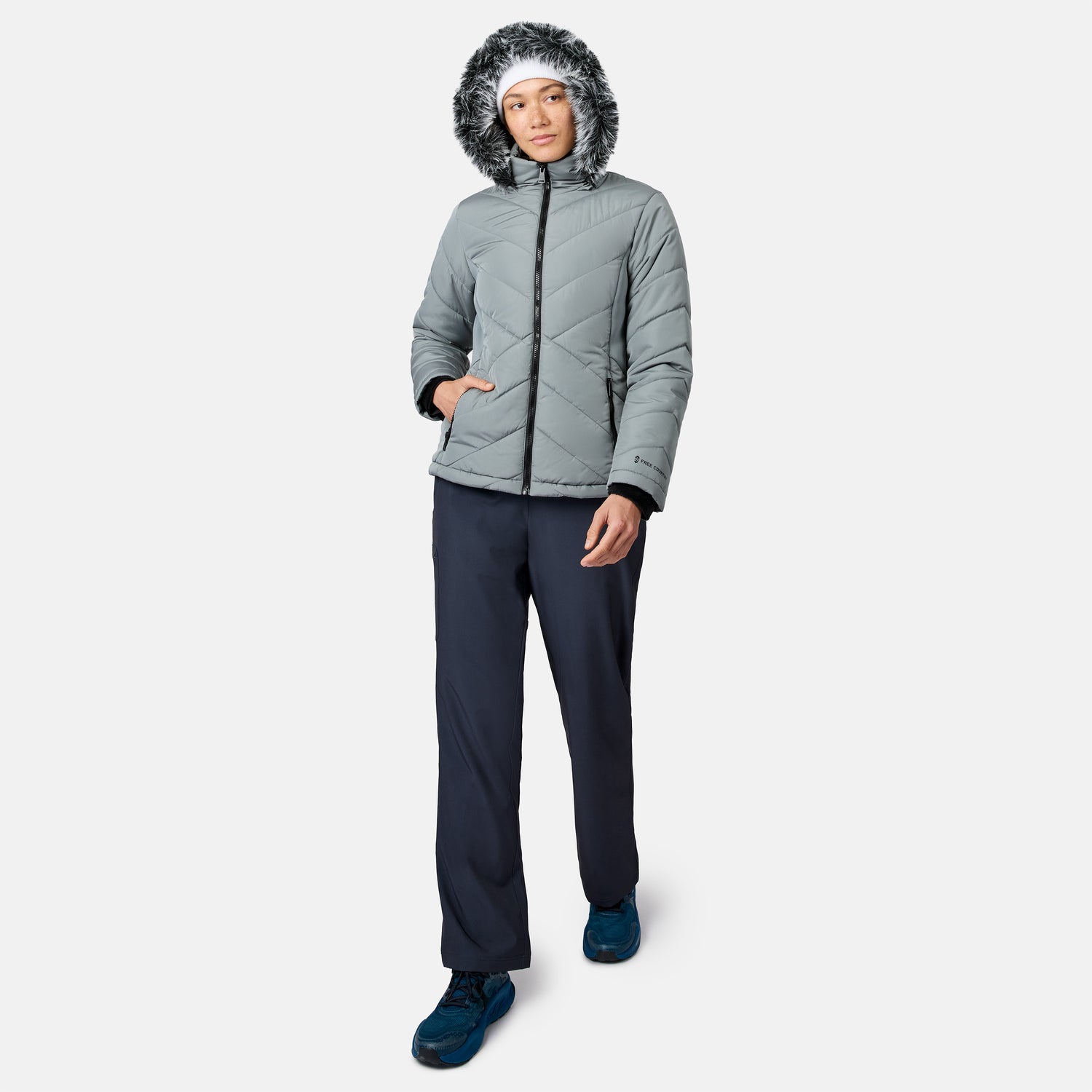 Person wearing a gray hooded puffer jacket with faux fur trim, Free Country Women’s Get Out There Fleece Lined Pants, and blue shoes, standing and facing forward against a plain white background.