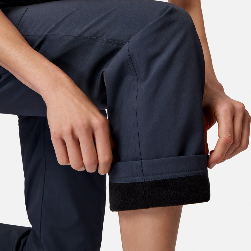 A person rolls up the cuff of Free Country Women’s Get Out There Fleece Lined Pants in dark blue, revealing a black fleece lining. Only their hands and part of their legs show against a plain background.