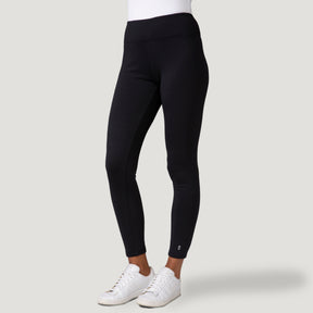 A person models Free Country Women's Force Grid Fleece Base Layer Bottoms, showcasing the black high-waisted leggings with four-way stretch and white sneakers against a plain light gray background; only their lower half is visible.