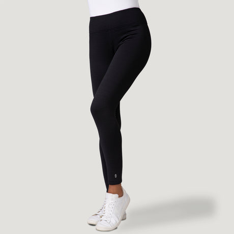 A person is shown from the waist down wearing Free Country Women's Force Grid Fleece Base Layer Bottoms in black, paired with a white top and white sneakers, standing against a light gray background.