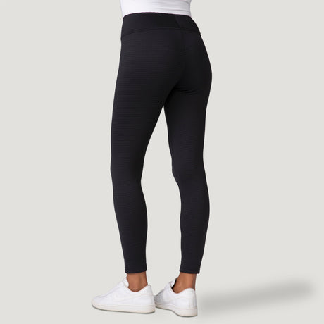 A person wearing Free Country Women's Force Grid Fleece Base Layer Bottoms is standing against a plain light gray background, shown from the back with white sneakers and their upper body partially visible.