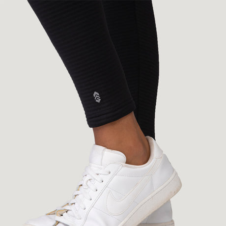 Close-up of a person wearing Free Country Women's Force Grid Fleece Base Layer Bottoms with four-way stretch and a small white logo near the ankle, paired with white Nike sneakers against a plain, light-colored background.