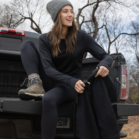 A woman in a gray knit beanie, black long-sleeve top, Free Country Women's Force Grid Fleece Base Layer Bottom leggings, and hiking boots sits on an open pickup tailgate, smiling with bare trees behind her.