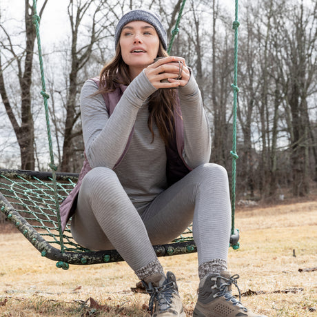 A woman in a gray beanie, patterned socks, hiking boots, and Free Country Women's Force Grid Fleece Base Layer Bottom sits on a green rope swing, holding a mug outdoors on a cloudy day with bare trees behind her.