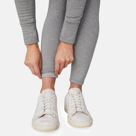 A person wearing Free Country Women's Force Grid Fleece Base Layer Bottom in light gray with white sneakers is shown adjusting the hem of one leg against a plain white background.