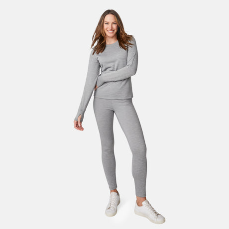 A smiling woman models the Free Country Women's Force Grid Fleece Base Layer Bottom in light gray, paired with a matching long-sleeve top and white sneakers against a plain white background. One arm is bent while her other hand rests by her side.