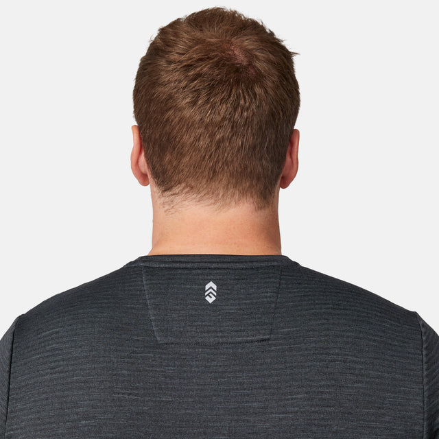 A person with short, light brown hair is seen from behind, wearing the Free Country Men's Force Grid Fleece Base Layer Top in dark gray, which features a small white chevron logo on the upper back, set against a plain light background.