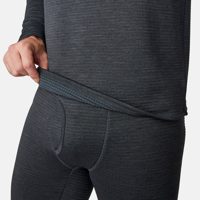 A person wearing the Free Country Men's Force Grid Fleece Base Layer Top in dark gray pulls up the top to reveal the waistband and textured fabric, with a plain white background.