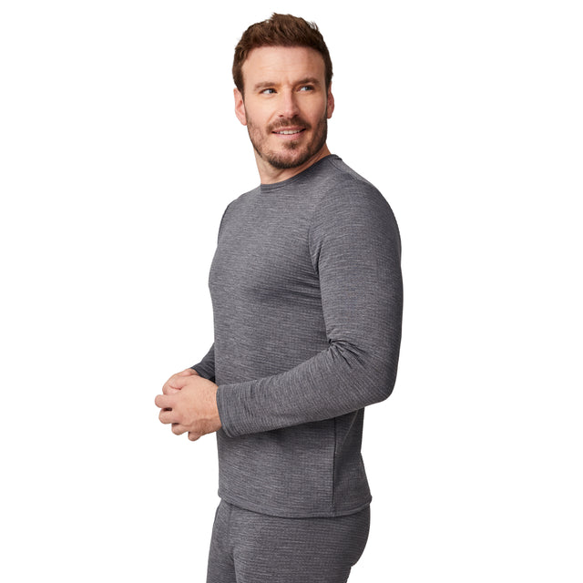 A man stands sideways, looking to his left with his hands loosely clasped, wearing the Free Country Men's Force Grid Fleece Base Layer Top in gray. The background is plain white.