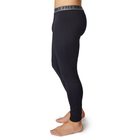 A barefoot man, shown from the chest down, wears the Free Country Men's Force Grid Fleece Base Layer Bottom—dark fitted leggings with four-way stretch and a gray waistband featuring FREE COUNTRY in black letters.