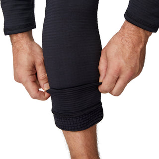 A person wears Free Country’s Men's Force Grid Fleece Base Layer Bottom in black, showing off the ribbed, textured fabric and stretchy fit while pulling up one leg; their hands and forearms are also visible.