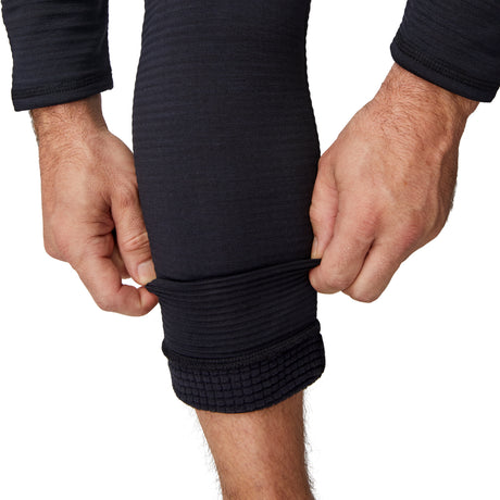 A person wears Free Country’s Men's Force Grid Fleece Base Layer Bottom in black, showing off the ribbed, textured fabric and stretchy fit while pulling up one leg; their hands and forearms are also visible.