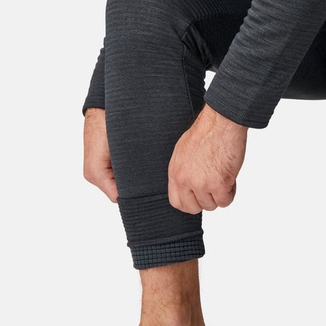 A person adjusts the cuff of their Men's Force Grid Fleece Base Layer Bottom from Free Country, showing a close-up of their hands and lower legs in dark gray ribbed thermal leggings against a plain white background.
