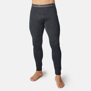 A man stands barefoot on a white background wearing Free Country's Men's Force Grid Fleece Base Layer Bottom in dark gray, featuring a visible elastic waistband and moisture-wicking fleece fabric.