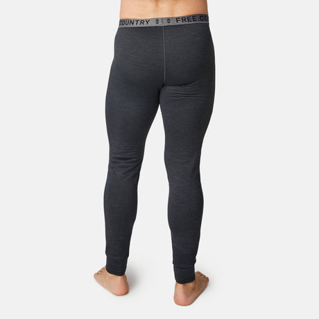 A barefoot person faces away, wearing Free Country Men's Force Grid Fleece Base Layer Bottoms in dark gray with ribbed texture and four-way stretch. The waistband shows the brand name. Photo is on a plain white background.