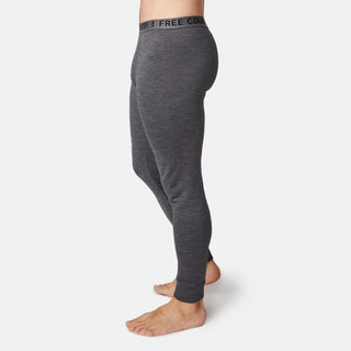 A man stands barefoot in Free Country’s Men's Force Grid Fleece Base Layer Bottoms—gray, fitted leggings with a black FREE COUNTRY waistband and four-way stretch—shown from the side on a plain white background.