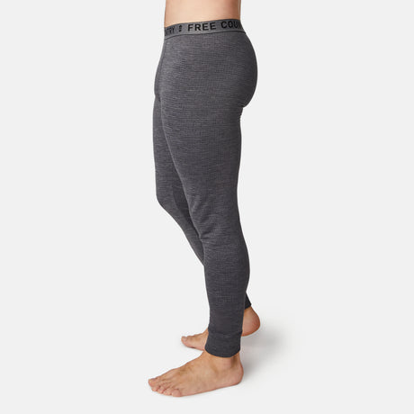 A man stands barefoot in Free Country’s Men's Force Grid Fleece Base Layer Bottoms—gray, fitted leggings with a black FREE COUNTRY waistband and four-way stretch—shown from the side on a plain white background.