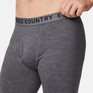 A man wears close-fitting heather gray Free Country Men's Force Grid Fleece Base Layer Bottoms, made from four-way stretch fleece, pulling the branded waistband outward with one hand.