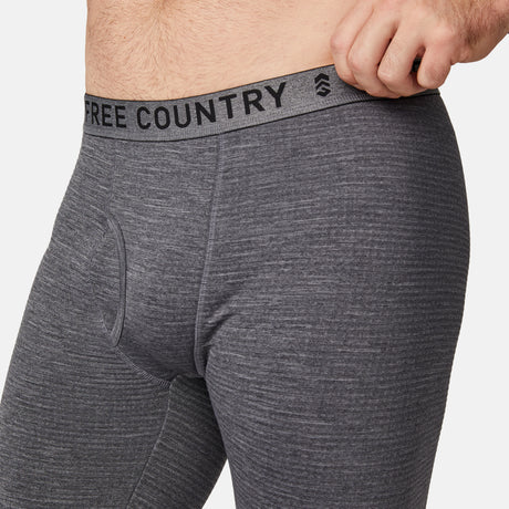 A man wears close-fitting heather gray Free Country Men's Force Grid Fleece Base Layer Bottoms, made from four-way stretch fleece, pulling the branded waistband outward with one hand.